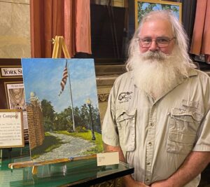A man poses, smiling, next to a painting. The painting is on a table, and it is propped up by a small wooden easel.